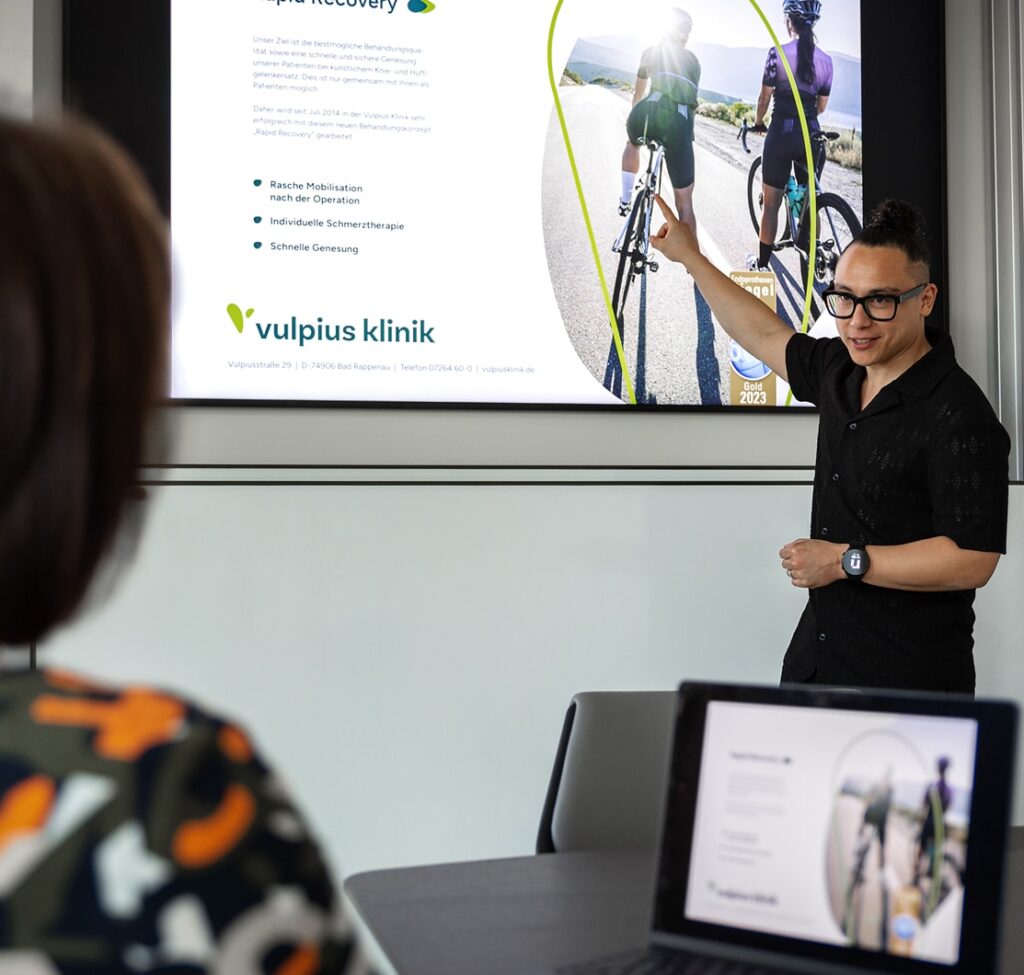 Harry Heckmann präsentiert Folie der Vulpius Klinik in Besprechungsraum; Teilnehmerin im Vordergrund.